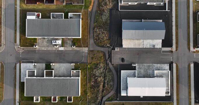 Top-down drone view in Grindavik, Iceland, showing a long fracture zone beside homes, splitting asphalt and rocky ground through a residential block after volcanic unrest.