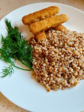 Cooked Buckwheat with Fish Sticks and Fresh Dill on a Plate. A serving of cooked buckwheat groats with crispy breaded fish sticks and fresh dill arranged on a white plate. Simple homemade meal combini