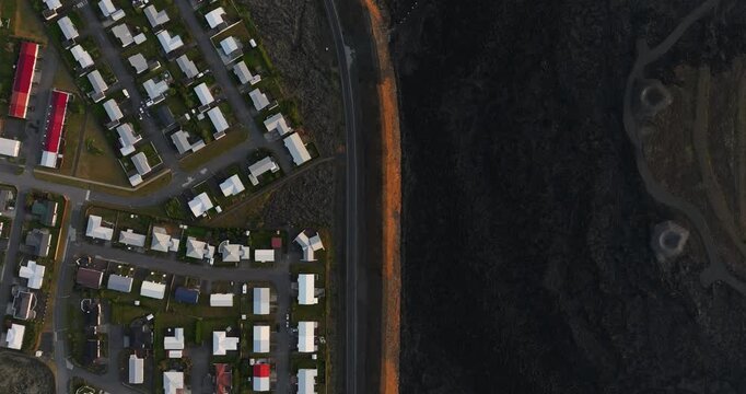 Top-down drone view of Grindavik, Iceland, with dense housing blocks beside a road and orange defense berm sharply dividing the town from an immense hardened black lava field.