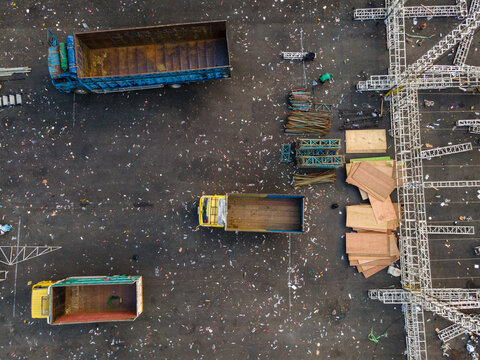 Aerial view of three dump trucks parked in an open lot with scattered construction materials, metal trusses, and wooden boards in Purwokerto, Central Java, Indonesia.