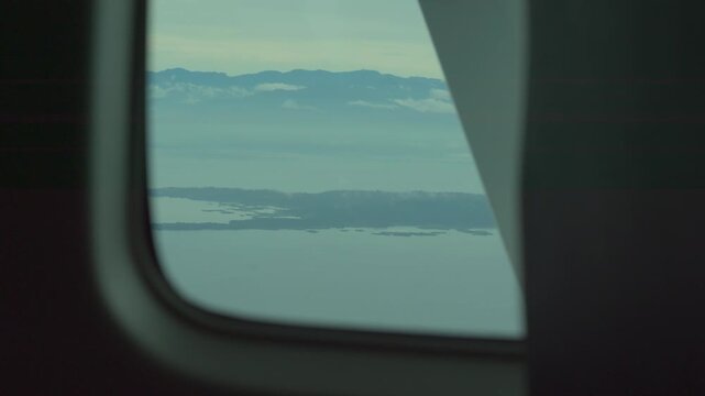 Airplane window view shows calm sea and distant islands. Clouds drift across the sky over the water. Coastal landforms become clearer through the frame. Peaceful perspective from an airplane journey