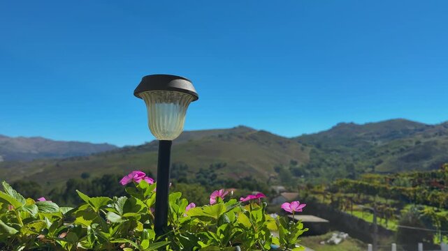 a classic solar-powered pathway light nestled among vibrant pink periwinkle flowers, overlooking a sunny rolling mountain valley.