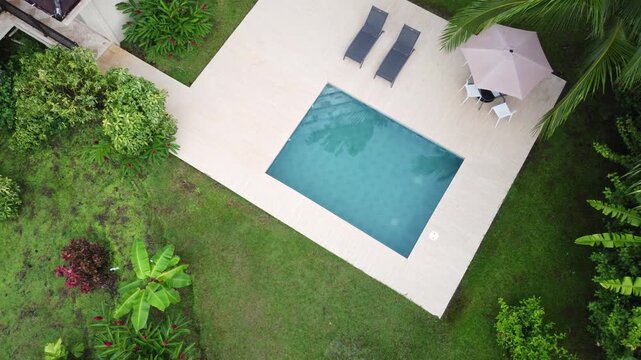 Aerial view of rectangular pool with clear blue water. Two lounge chairs and large umbrella on stone patio. Lush banana plants and palm trees surround the area