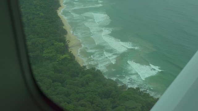 Airplane window reveals coastal view. Ocean waves crash onto sandy beach. Lush green forest lines the water's edge. Light clouds float above the sea. Ideal for travel or nature projects