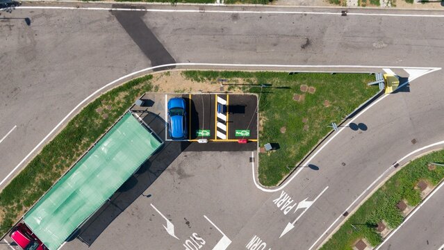 Aerial view of an electric vehicle charging station with a blue car parked in a designated spot, featuring road markings and a green canopy in Milan, Lombardy, Italy.