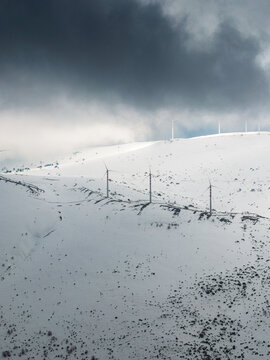 Aerial view of wind turbines standing on snow-covered mountain slopes under a dramatic dark cloudy sky in Anversa degli Abruzzi, Abruzzo, Italy.