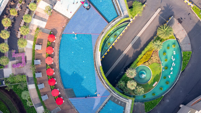 Aerial view of a modern resort swimming pool with vibrant red umbrellas, wooden decking, and lush landscaped gardens in Yogyakarta, Special Region of Yogyakarta, Indonesia.