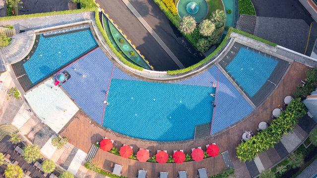 Aerial view of a curved swimming pool with blue water, red umbrellas, and lounge chairs on a wooden deck in Yogyakarta, Special Region of Yogyakarta, Indonesia.
