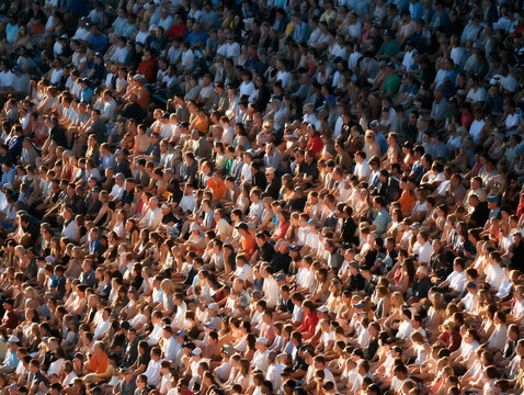 Large crowd of people sitting in a stadium, enjoying a live event in warm sunlight, showing diversity and unity among spectators.