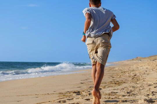 Teenage boy jogging barefoot along a sandy beach beside ocean