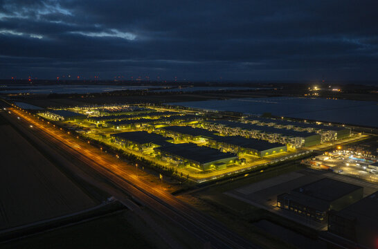 Aerial view of a large illuminated hyperscale AI data center complex at night, surrounded by dark agricultural fields and a highway in the Netherlands.