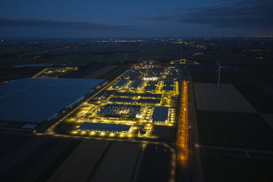 Aerial view of a hyperscale AI data center illuminated at night, featuring rows of server buildings, cooling infrastructure, and surrounding agricultural fields in the Netherlands.