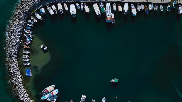 Overhead drone shot of a curved stone breakwater marina packed with small fishing boats on dark green water.