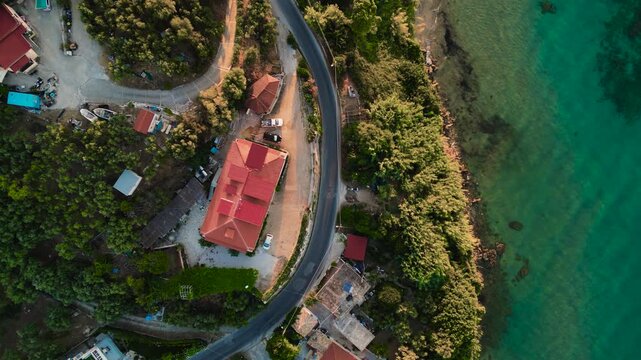Overhead drone shot of winding coastal road beside turquoise sea, red-roofed houses surrounded by lush trees.