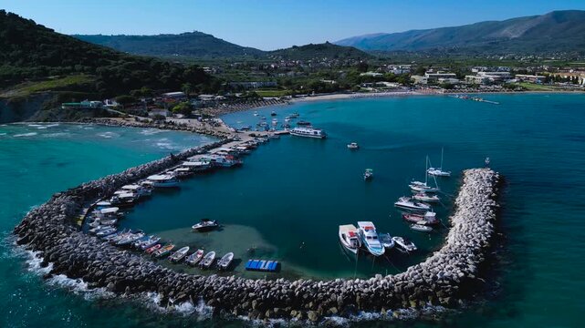Drone shot over Alykanas marina showing boats docked inside stone breakwater, turquoise Ionian Sea, sandy beach and green hills.