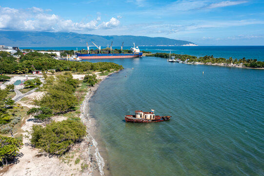 Aerial view of a rusted shipwreck in the coastal waters near the Barahona industrial port, with a large cargo ship docked in the background, Barahona, Barahona Province, Dominican Republic.