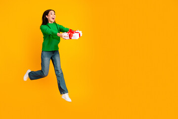 Naklejka premium Happy young woman jumping and offering a wrapped gift with red ribbon on a bright yellow background, joyful surprise and festive celebration of giving