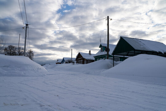 Snow blankets the village street. Snowdrifts line the road. Wooden houses stand under a cloudy sky. Power lines stretch across the scene. This quiet winter view suits travel or seasonal projects