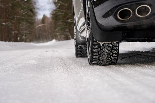 Black car's rear tires grip snowy road. Dual exhaust pipes gleam in winter light. Snow-covered path winds through forest trees. Winter driving safety concept shown