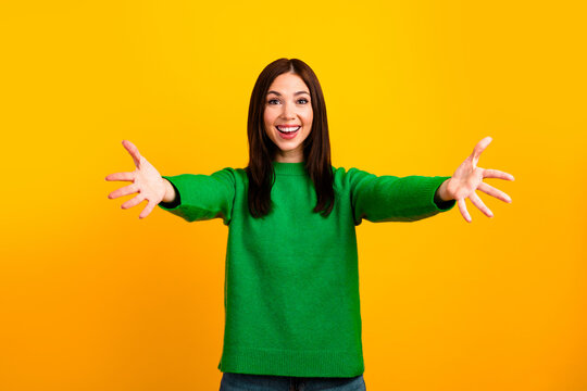 Smiling woman in green sweater with open arms against bright yellow background welcoming and offering a friendly hug, cheerful casual portrait expressing happiness and warmth