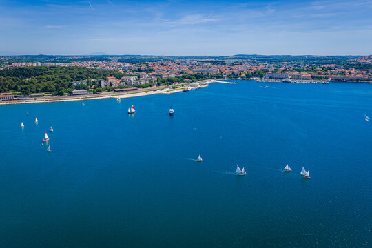 Aerial view of sailboats navigating the blue waters of the bay with the historic cityscape of Pula in the background under a clear sky Pula, Istria County, Croatia.