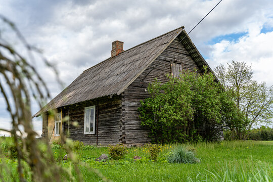 View of a weathered wooden house with a gabled roof set in a lush green field under a cloudy sky in Latvia.
