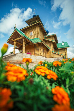 View of a traditional wooden church with green roof and blue domes, framed by vibrant orange marigold flowers in the foreground under a bright blue sky in Balvi, Latvia.