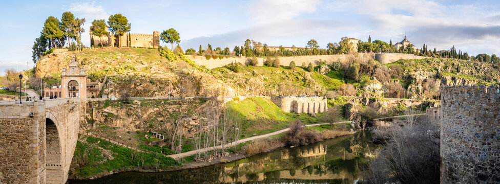 View of the historic Alcantara Bridge and ancient stone city walls overlooking the Tagus River in Toledo, Castile-La Mancha, Spain.