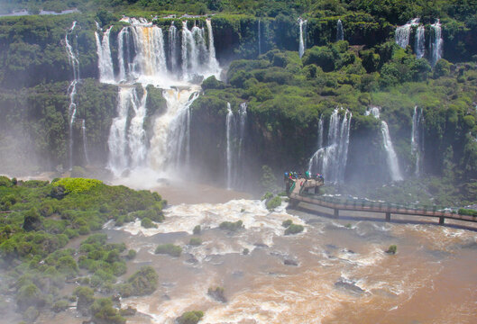 View of Iguazu Falls with cascading water, lush green vegetation, and a pedestrian walkway with people in Foz do Iguacu, Parana, Brazil.