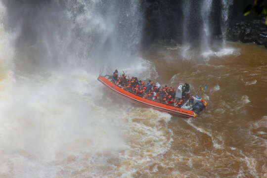 View of a tourist boat navigating the turbulent waters at the base of the powerful Iguazu Falls with passengers wearing orange life vests in Foz do Iguacu, Parana, Brazil.