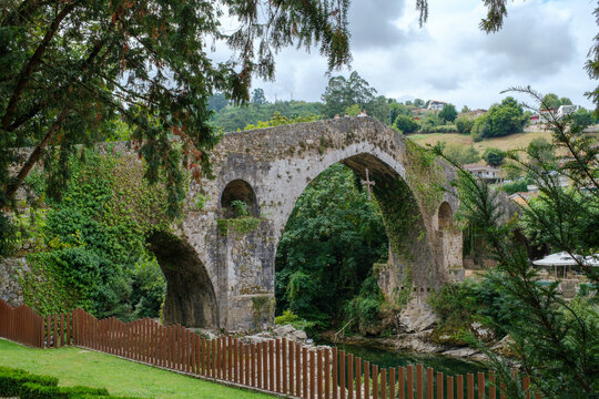 View of the historic Roman bridge in Cangas de Onis with its iconic hanging cross, surrounded by lush green trees and vegetation over the Sella River in Spain.