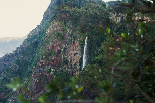 View of a tall waterfall cascading down a steep, lush green cliff face in the mountains of Madeira, Portugal.