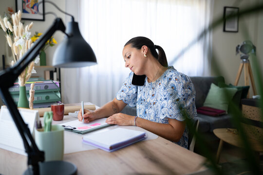 White woman multitasking with phone and notepad in home office setting