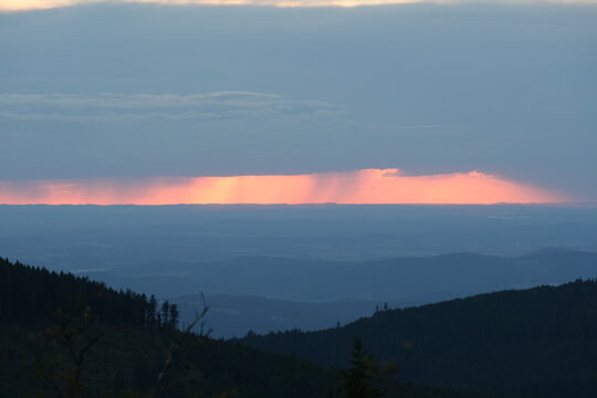 Dramatischer Sonnenuntergang von einem Berggipfel aus gesehen mit h&uuml;geliger Waldlandschaft in zarten Farbabstufungen und glutroter Sonne unter einem dunklen Wolkenband