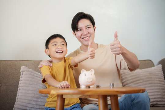 Asian father and son giving thumbs up beside piggy bank at home for financial literacy