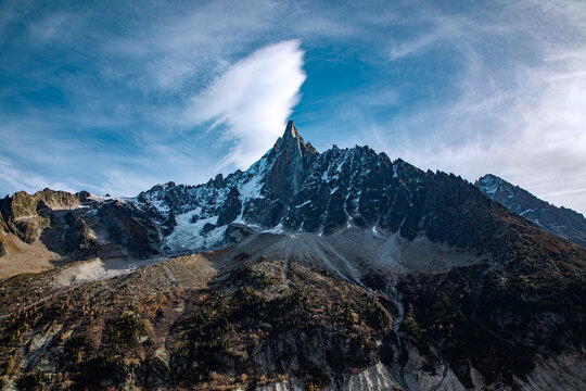 View of the majestic snow-capped Mont Blanc peak under a blue sky with a lenticular cloud over the summit, rugged rocky slopes and alpine terrain in Chamonix, Auvergne-Rhone-Alpes, France.