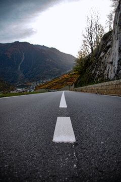 View of an asphalt road leading towards the Mont Blanc massif, with autumn-colored slopes, a stone retaining wall, and a cloudy sky in Chamonix, Auvergne-Rhone-Alpes, France.