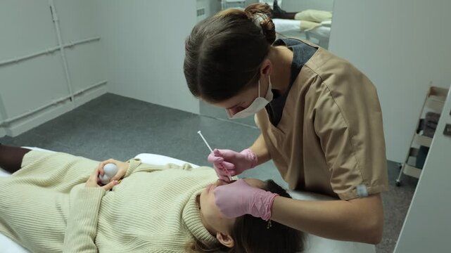A masked cosmetologist injects botox into a client's forehead with a syringe, seen in a wide shot of a modern beauty clinic room.