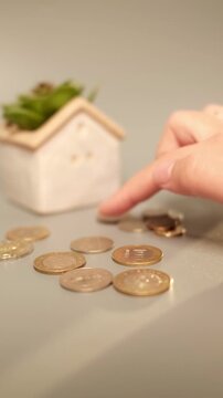 Kazakhstan tenge coins are moved one by one as if being counted beside a small house, creating a minimalist visual concept of saving money, home ownership, budgeting and financial planning.