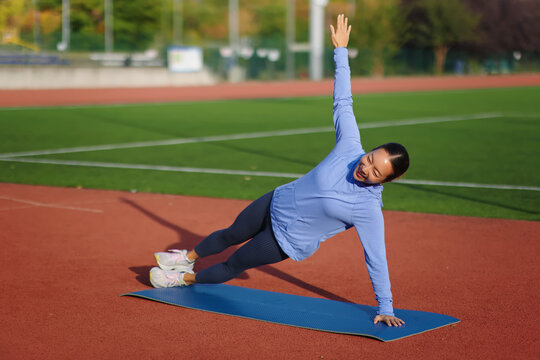 Young woman practicing side plank yoga pose on a blue mat, balancing on one arm and leg, exercising core strength and flexibility outdoors on a running track