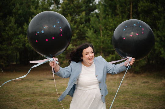 Joyful pregnant woman running and holding two black gender reveal balloons for twins outdoors in a forest.