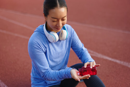 Young asian woman sitting on a running track taking supplements, holding a bottle and pouring red pills into her hand, prioritizing health and wellness in her athletic lifestyle