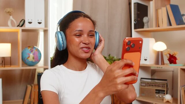 African American young woman on home table learns foreign language using smartphone and headphones and repeats new words. Black girl practices speaking and pronunciation during online study at home.