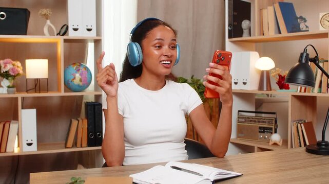 African American young woman on home table learns new words and repeats phrases during language lesson. Black girl studies with headphones smartphone and notebook feeling focused and engaged at home.