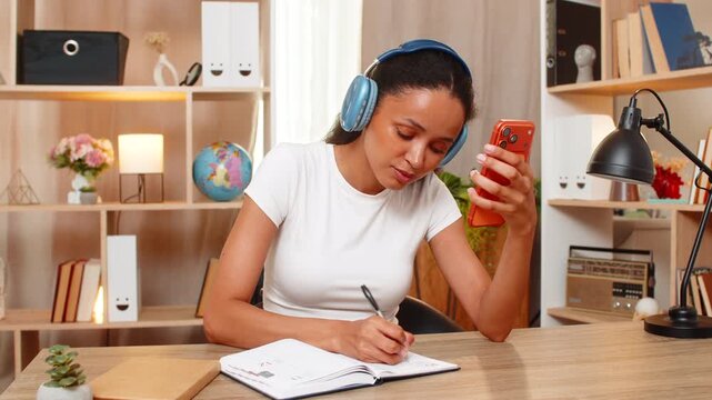 African American young woman on home table writes new foreign word in notebook during online language lesson on smartphone. Black girl studies words with headphones and builds speech skills at home.