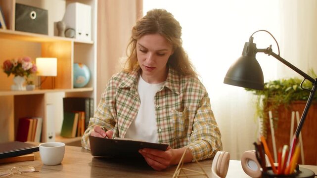 Young woman on home table reads contract on clipboard signs final page and feels pleased with work result. Adult girl on home table checks final paragraph then looks at camera with calm proud smile.