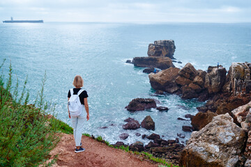 A woman with a white backpack stands on a rocky cliff edge overlooking the calm blue ocean and rugged coastline, conveying solitude, travel, adventure and peaceful seaside exploration. © Loginova