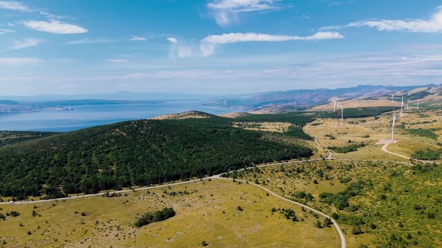 Aerial video overlooking a rugged coastal landscape where forested hills and golden mountain meadows descend toward the deep blue Adriatic Sea. Wind turbines are visible across the distant hills