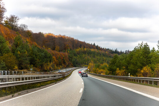 Autumn highway curves through colorful forest under cloudy sky, with cars traveling along open asphalt road. Seasonal foliage, transportation and scenic countryside create calm road trip atmosphere