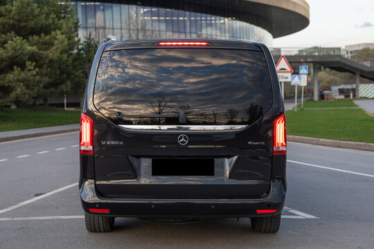 A premium black Mercedes-Benz V-Class minivan parked on a stylish city street during a sunny day. Modern urban skyline with glass skyscrapers in the background, professional executive shuttle, luxury 
